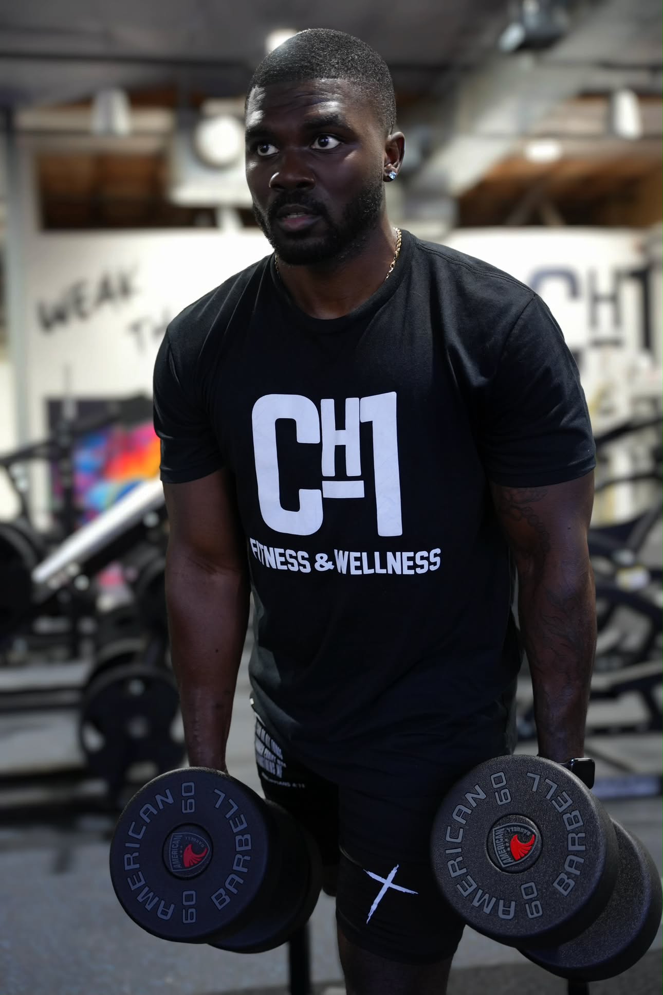 Jeremiah Myles holding two dumbbells in the gym, wearing a CH1 Fitness & Wellness shirt